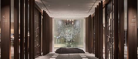 Un hall d’hôtel moderne avec un mur central en verre, des panneaux de bois et un sol à motifs.