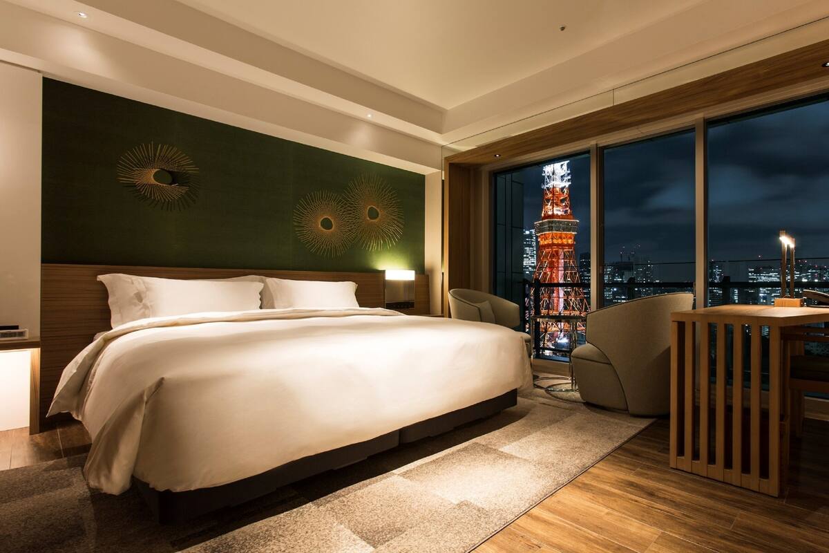 Une chambre d’hôtel moderne avec un grand lit, une vue sur la tour de Tokyo la nuit et un balcon équipé d’une table et d’une chaise.