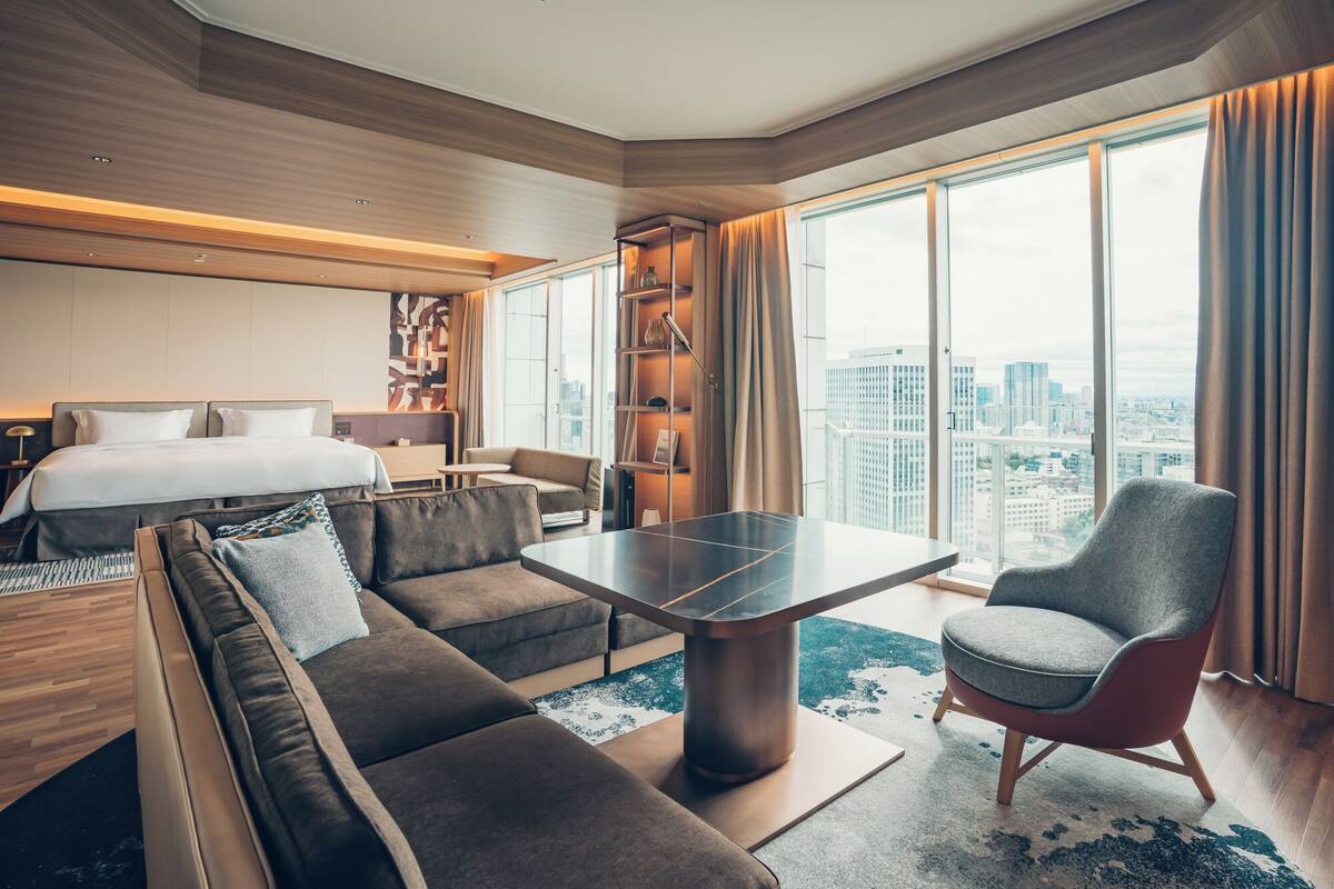 Une chambre d’hôtel moderne avec un grand lit, un canapé, une table basse et un fauteuil. On y trouve également une bibliothèque et une vue sur la ville grâce à de grandes fenêtres.