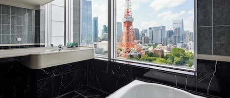 A modern bathroom with a large bathtub, a vanity with a mirror, and a window offering a view of a cityscape including a prominent tower.