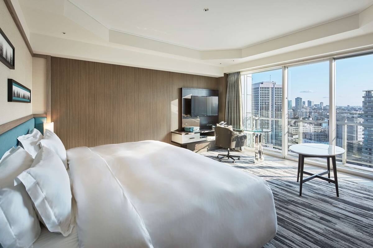Une chambre d’hôtel avec un grand lit, un bureau doté d’un téléviseur à écran plat, une chaise et une petite table. La chambre offre une vue sur la ville grâce à de grandes fenêtres.