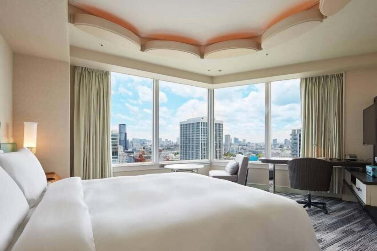 Une chambre d’hôtel avec un grand lit, un bureau, une chaise et une vue sur le paysage urbain grâce à de grandes fenêtres.