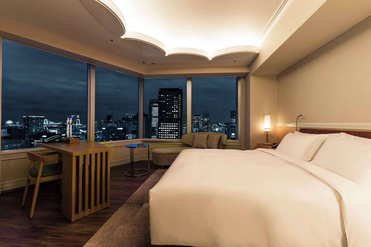 Une chambre d’hôtel avec un grand lit, un bureau, une chaise et une vue sur le paysage urbain la nuit.