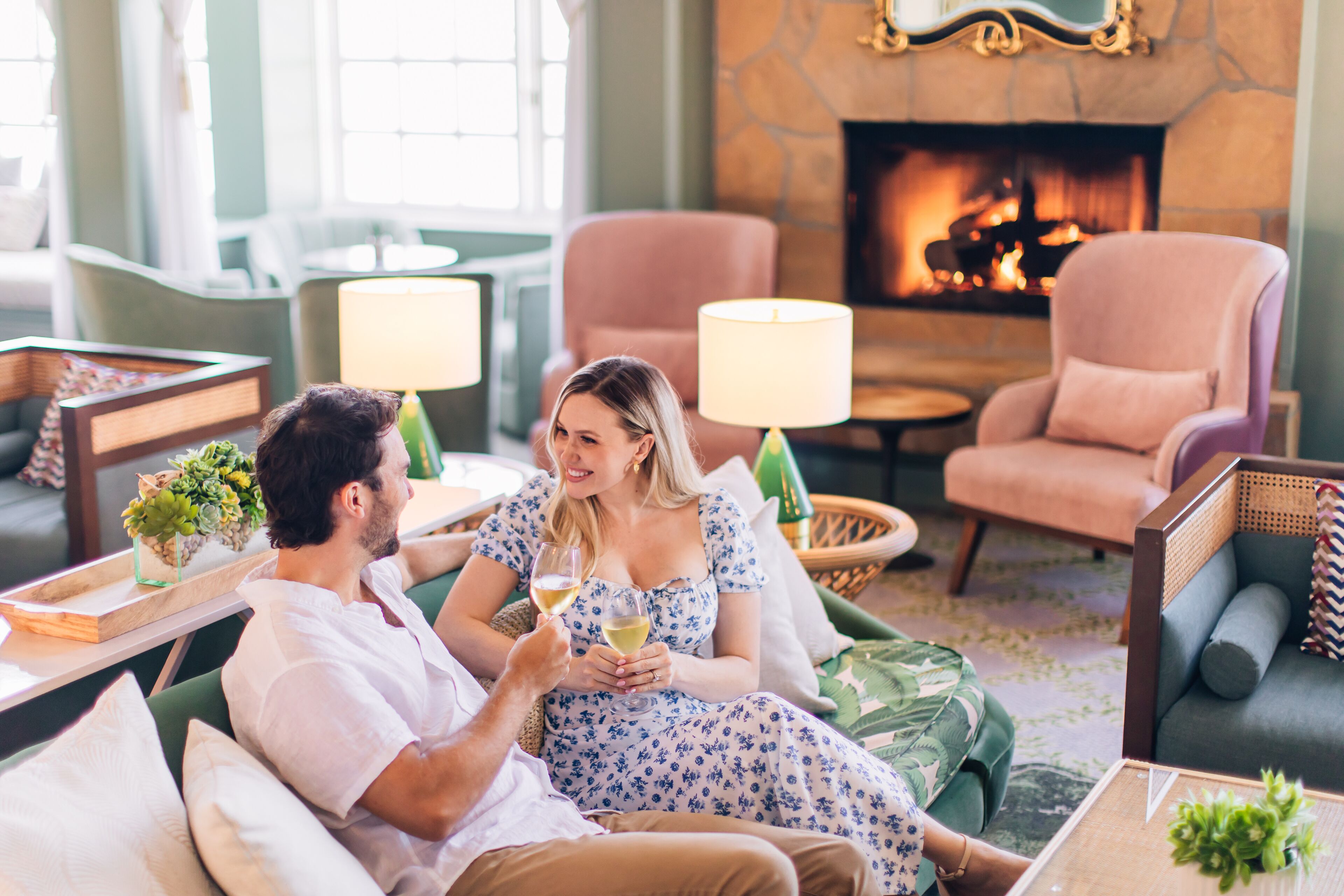 A couple seated on a sofa in a well-lit room with a fireplace, surrounded by comfortable seating and a table with a lamp.