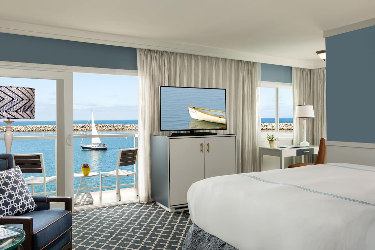 A hotel room with a large bed, a television displaying a boat on water, a desk with a lamp, and a view of the sea through a balcony.