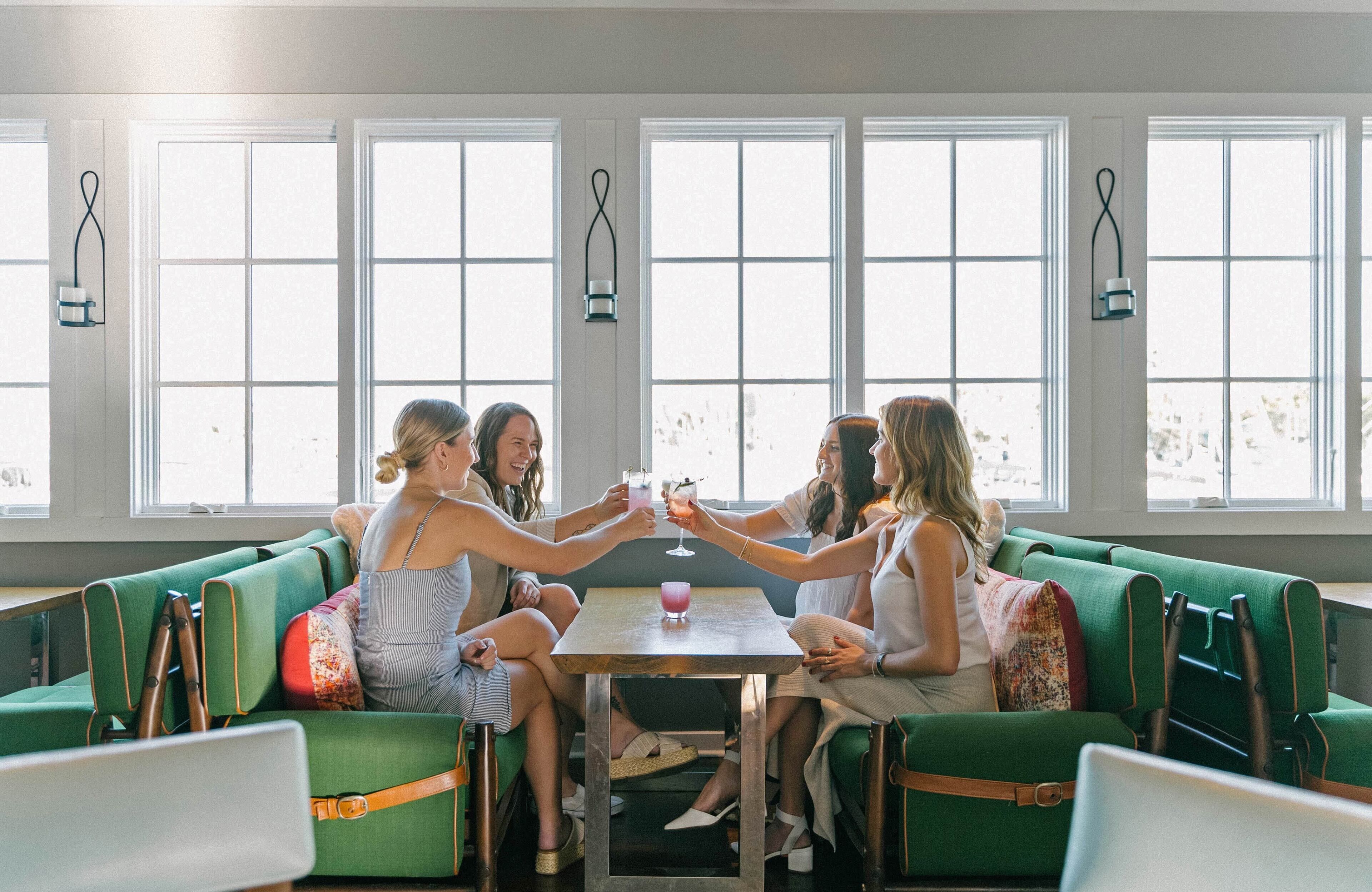 A group of people seated on green upholstered chairs around a wooden table in a room with large windows.