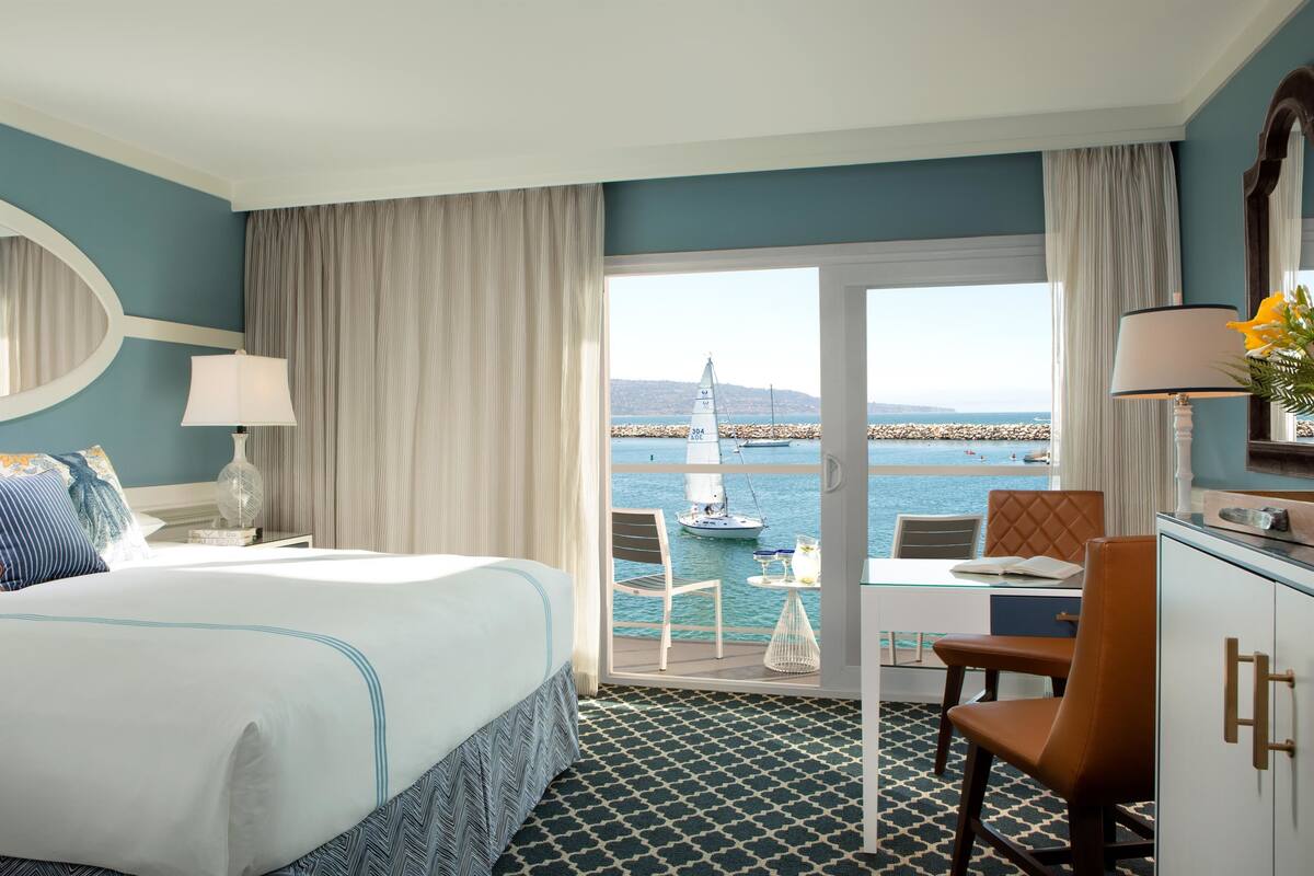 A hotel room with a bed, a desk, a chair, a lamp, and a view of the sea.