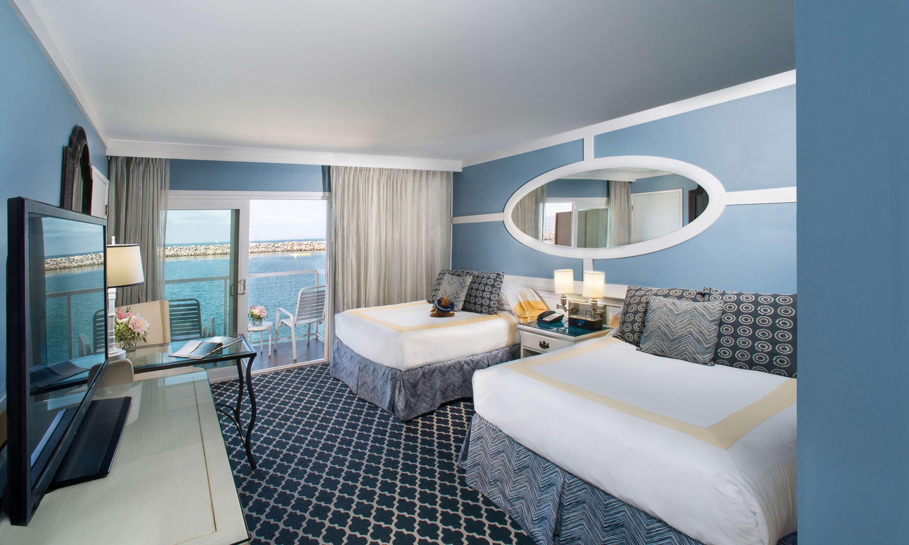 A hotel room with two beds, a desk with a lamp, a TV, a balcony with a view of the sea, and a large mirror.
