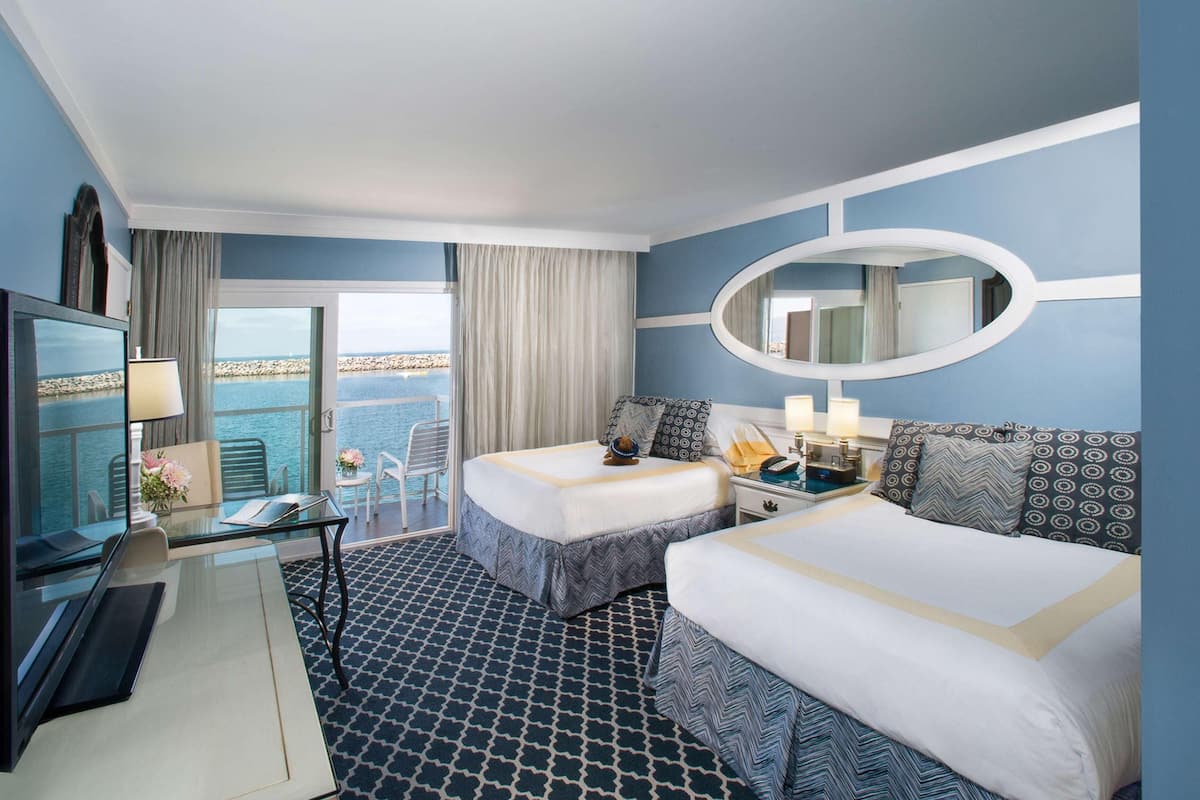 A hotel room with two beds, a desk with a lamp, a TV, a balcony with a view of the sea, and a large mirror.