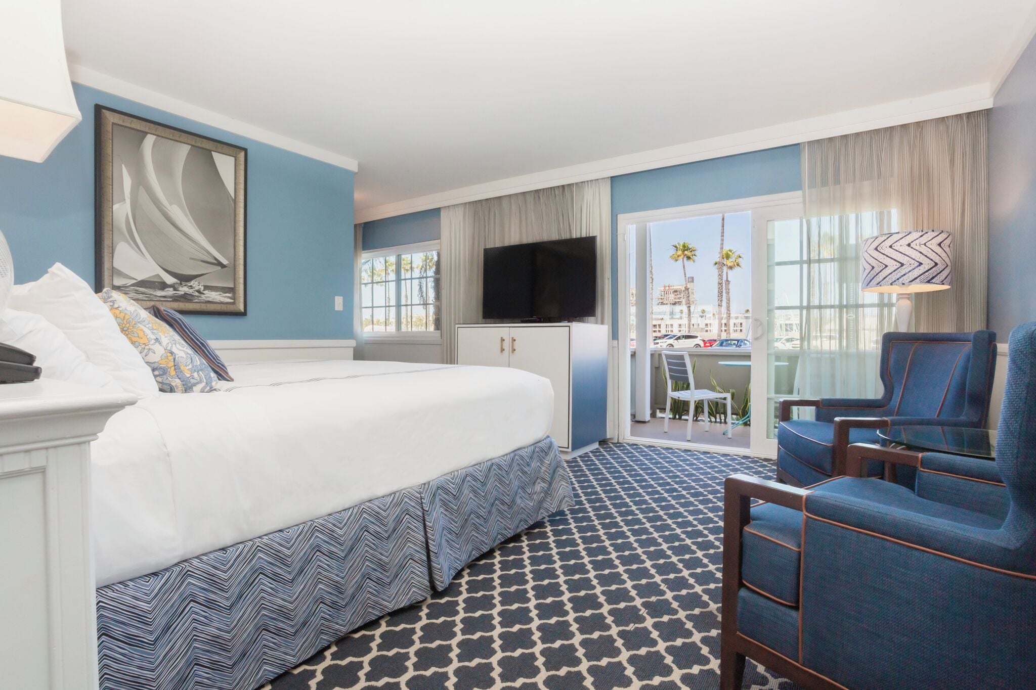 A hotel room with a bed, a TV, a seating area with chairs, a desk, and a view of palm trees.
