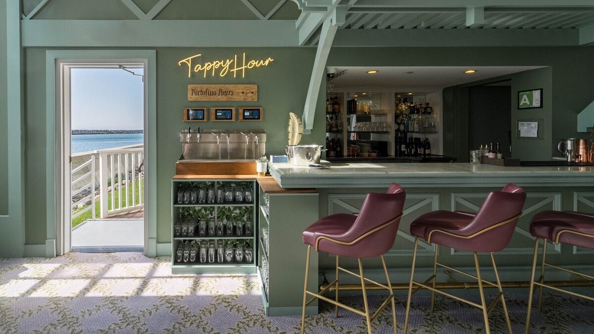 A bar area with a view of the sea, a neon sign reading "Happy Hour," and a shelf with glassware.