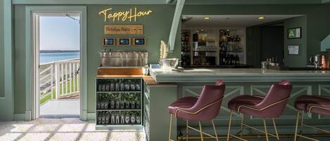 A bar area with a view of the sea, a neon sign reading "Happy Hour," and a shelf with glassware.