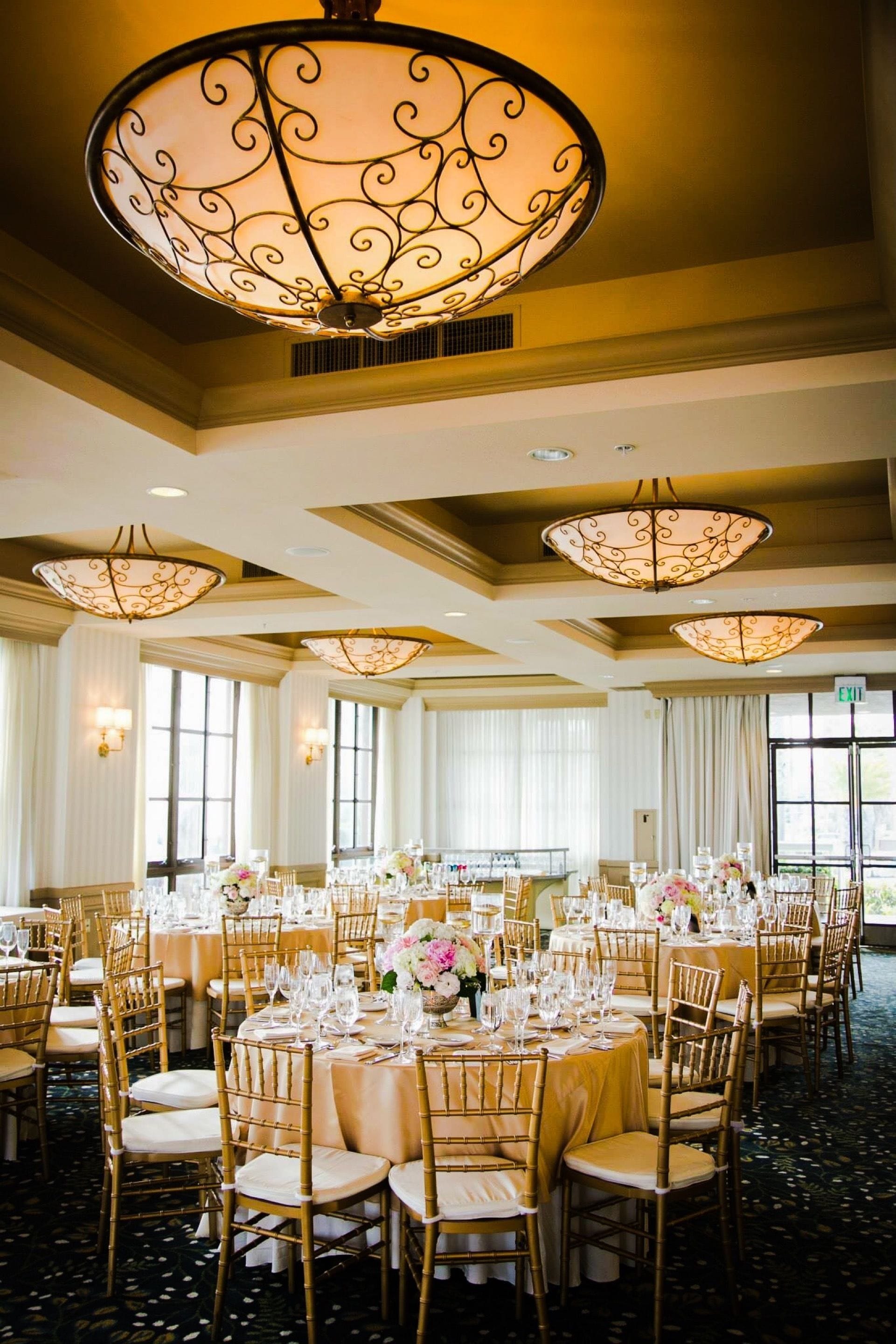 A banquet hall with round tables, gold chairs, and large chandeliers.