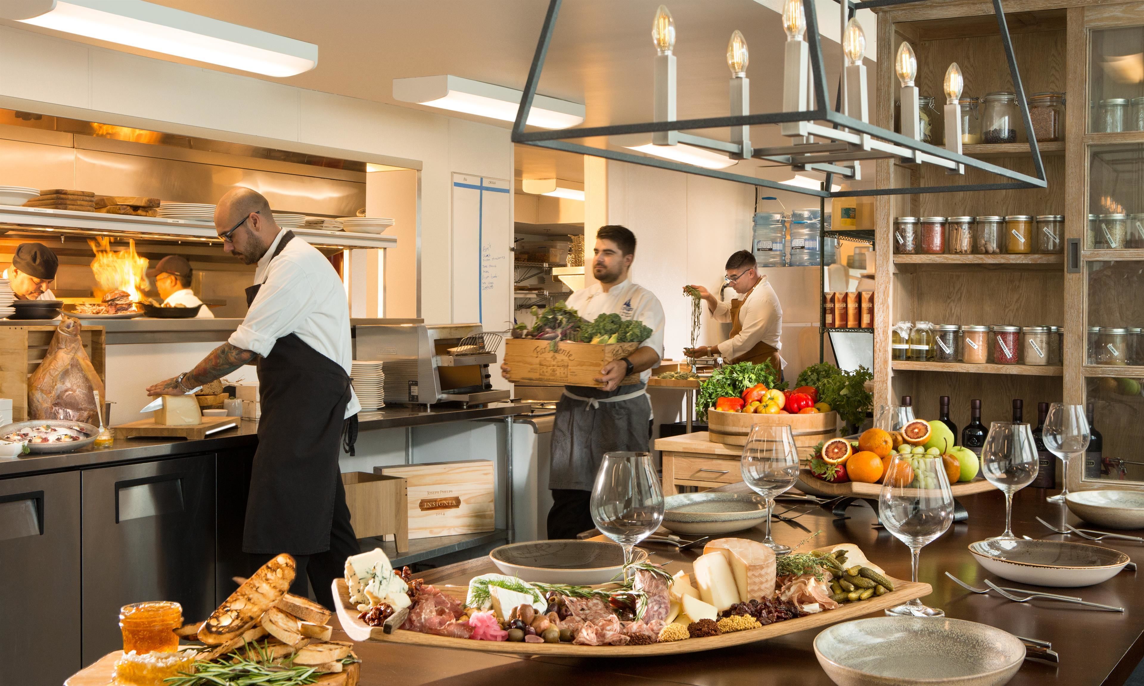 Una cucina ristorante moderna con chef che preparano cibo, un tavolo buffet con diversi piatti e bevande, e una dispensa con bottiglie di vino.