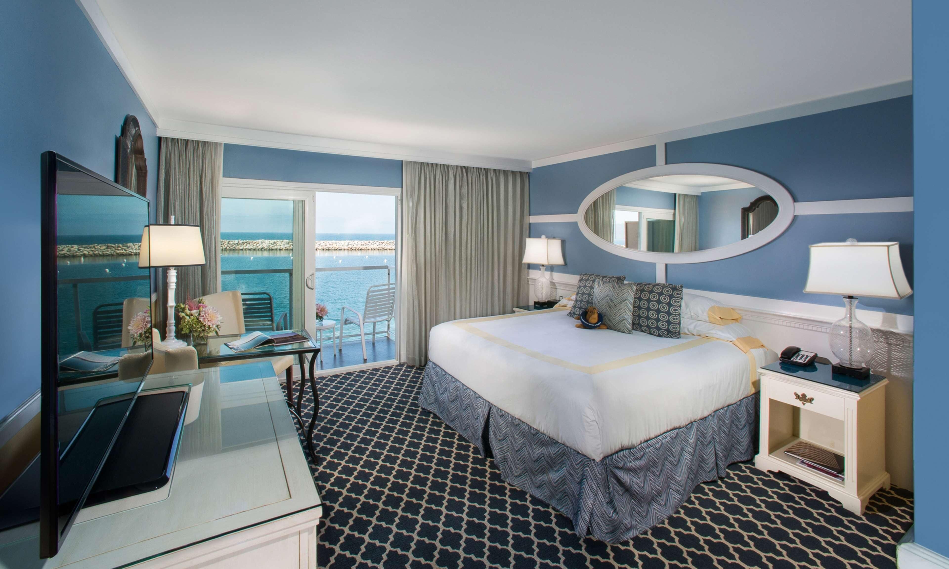 A hotel room with a bed, a desk with a laptop, a chair, a nightstand with a lamp, and a view of the sea.