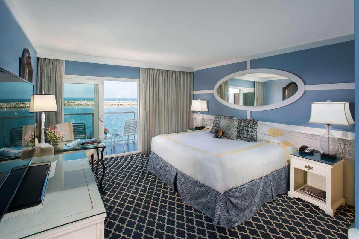 A hotel room with a bed, a desk with a laptop, a chair, a nightstand with a lamp, and a view of the sea.
