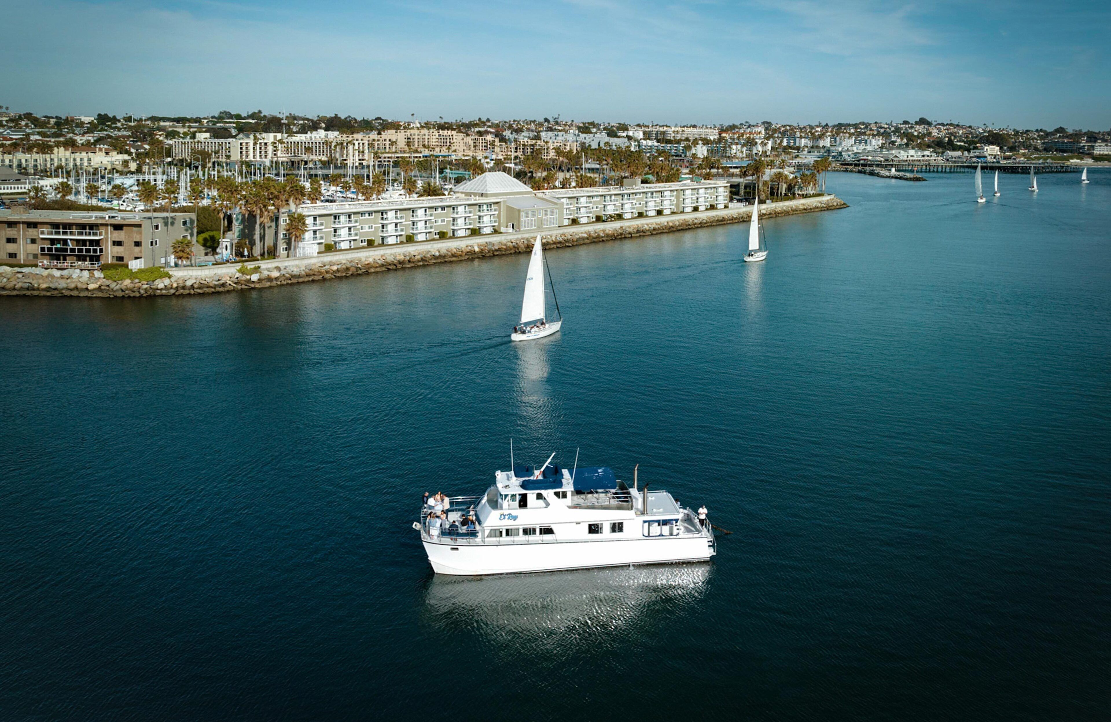 En lysthavn med en stor hvid yacht til anker, flere sejlbåde og boligbygninger i baggrunden.