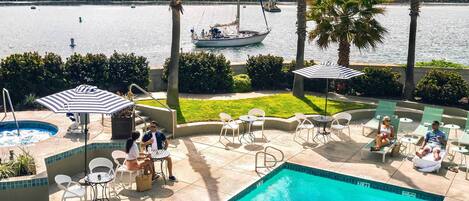 A pool area with lounge chairs and umbrellas, a view of the water with boats, and a stone wall in the background.