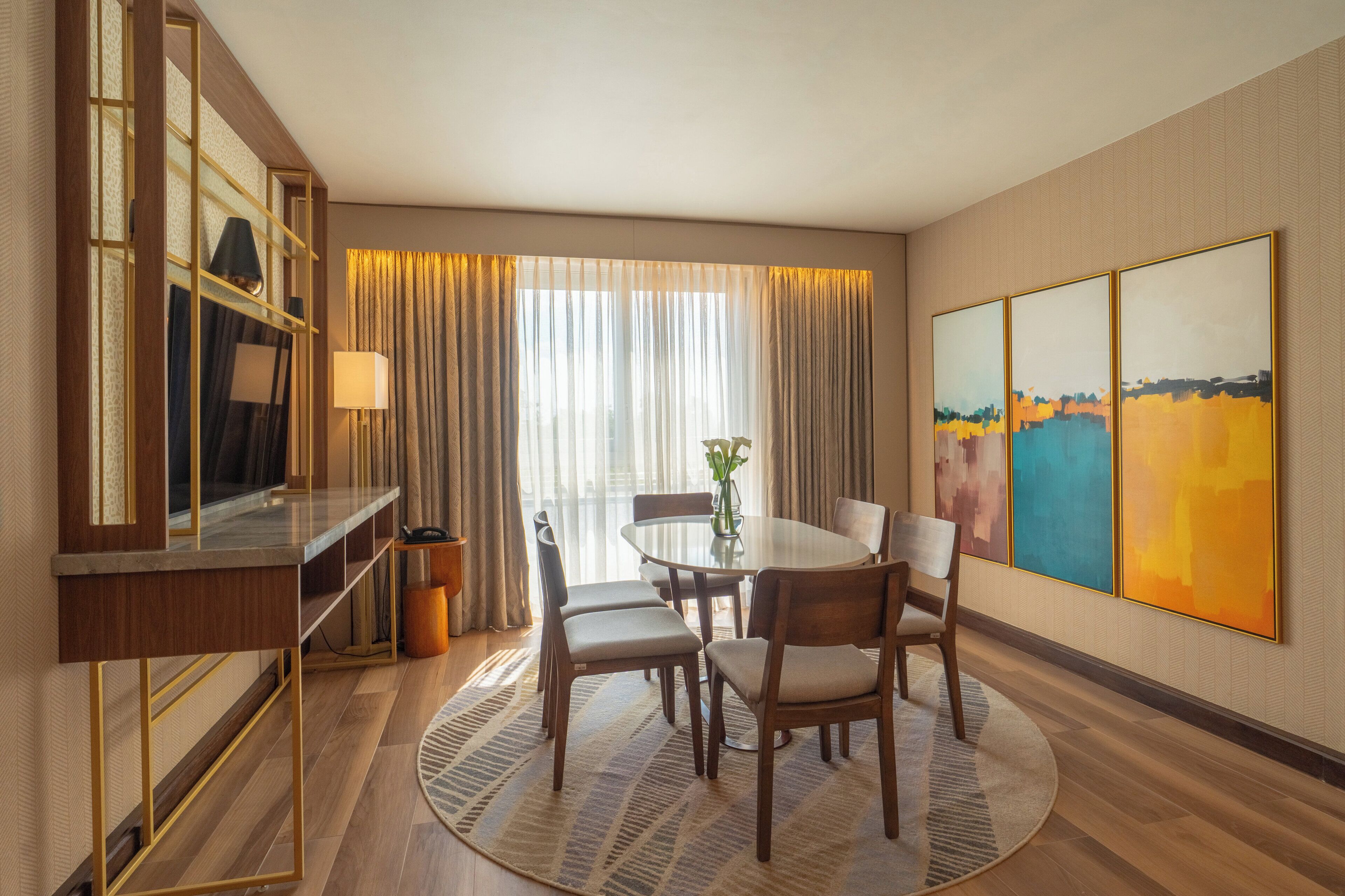 Ein Hotelzimmer mit Essbereich, einem runden Tisch und Stühlen. Ein Fenster mit Vorhängen, ein Bild an der Wand und ein kleiner Nachttisch mit Lampe.