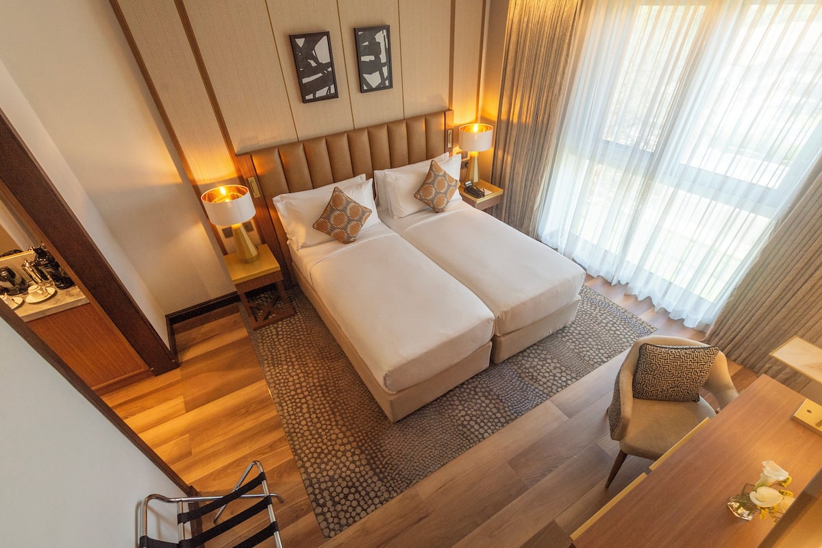 A hotel room with a double bed, two bedside tables with lamps, a chair, and a view of the outdoors through sheer curtains.