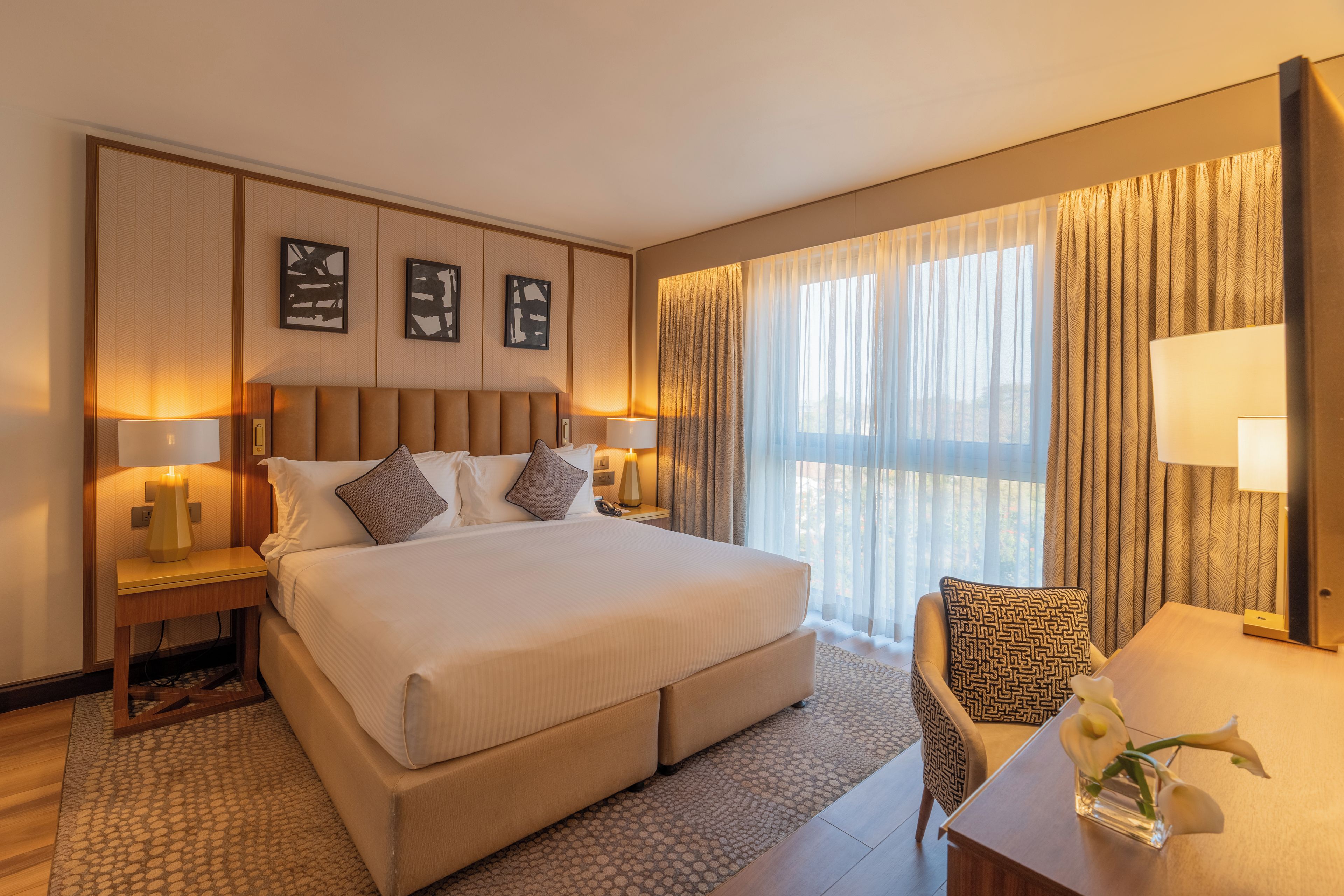 A hotel room with a large bed, two bedside tables with lamps, a chair, and a view of the outdoors through sheer curtains.