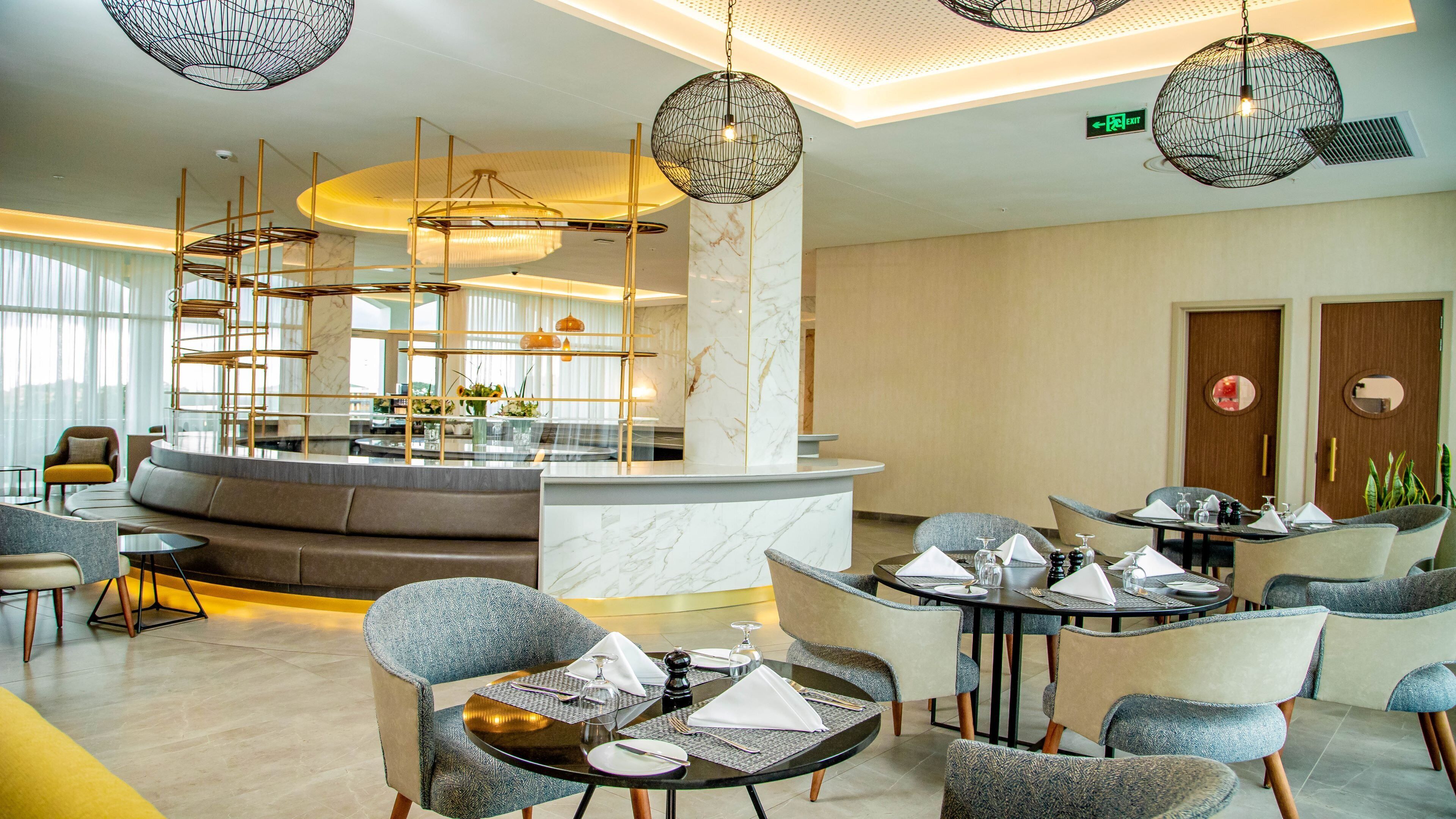 A modern hotel lobby with a central seating area, a reception desk, and a dining space.