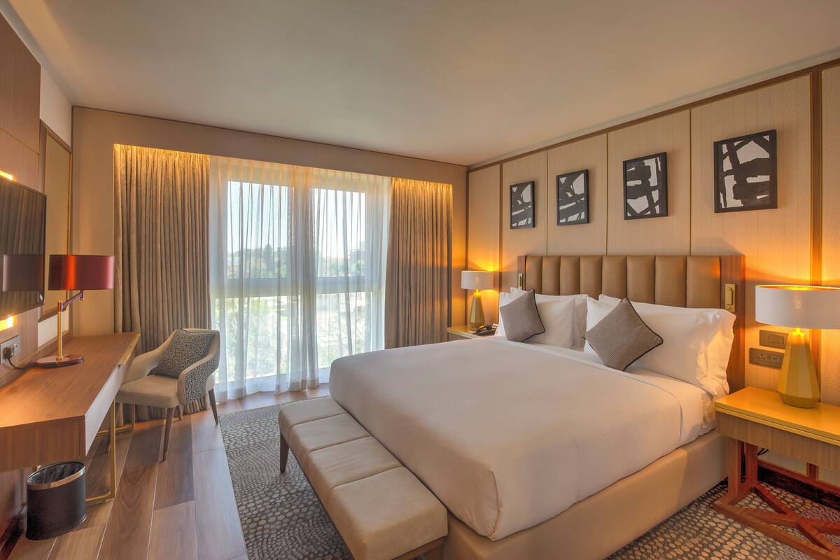 A hotel room with a large bed, a desk, a chair, and a view of the outdoors through the window.