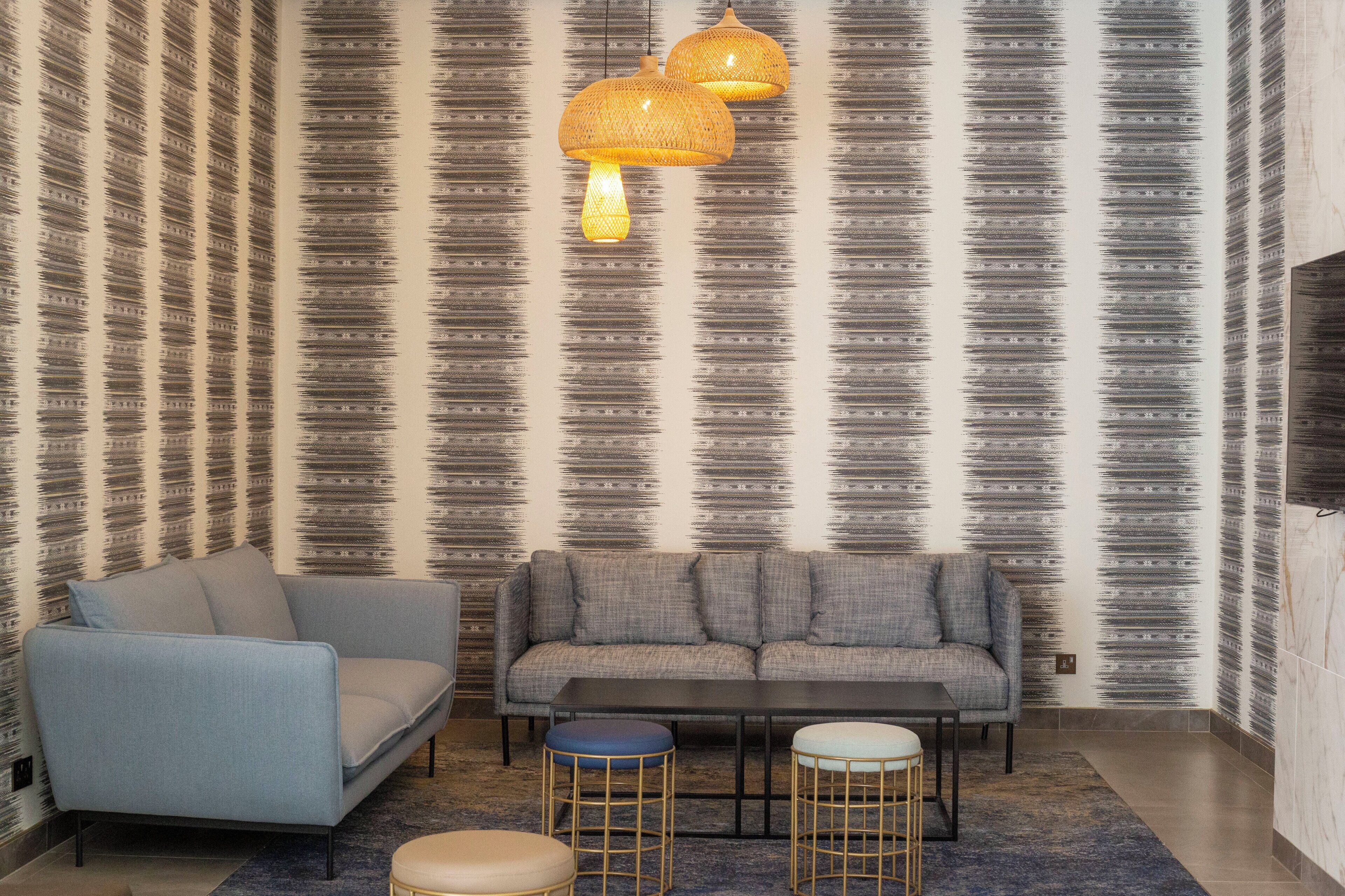 A modern living room with a patterned wallpaper, a sofa, a blue armchair, and two stools.