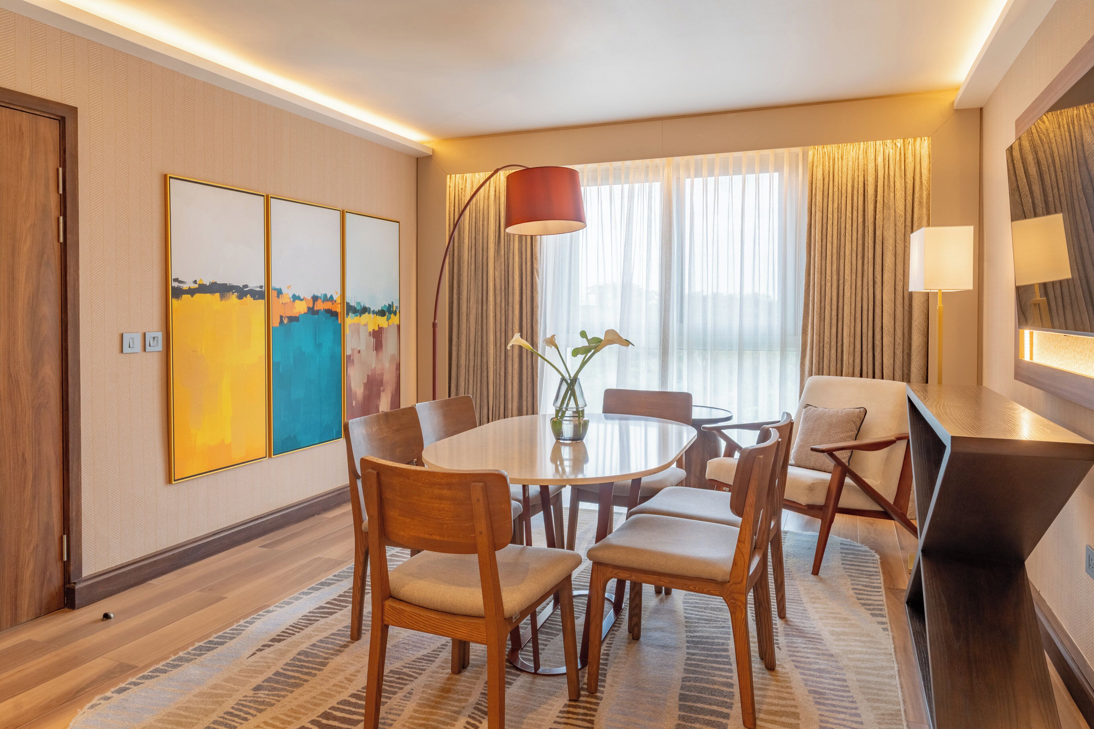 A hotel room with a round dining table, wooden chairs, a vase with flowers, and a large window with curtains.