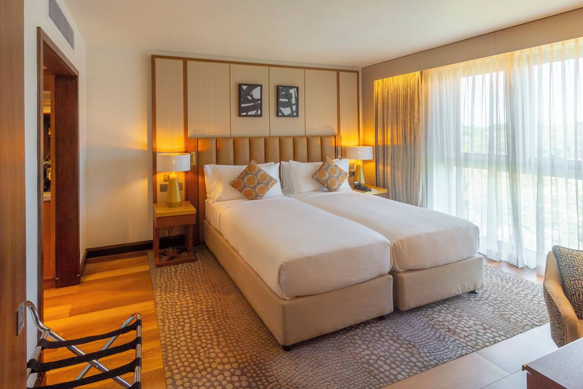 A double bed with two pillows, a bedside table with a lamp, and a window with sheer curtains.