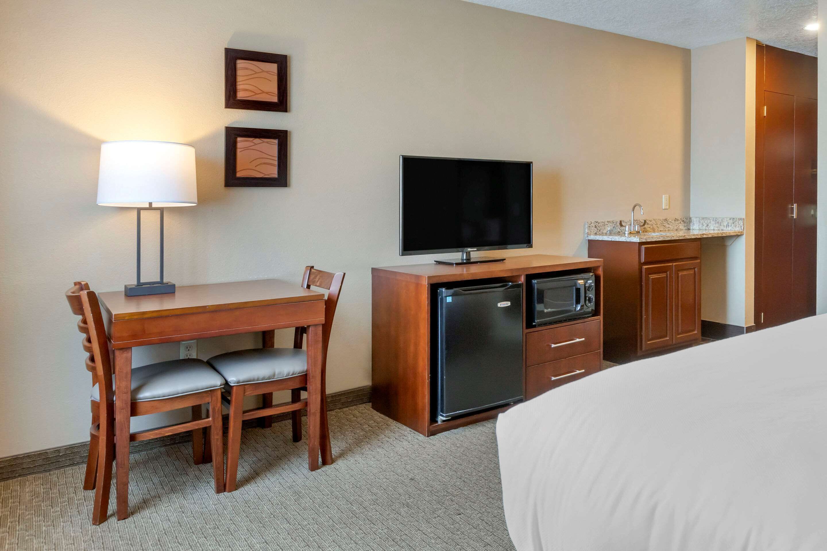 A hotel room with a bed, a desk with a lamp, a television, a refrigerator, a microwave, and a sink.