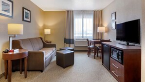 A hotel room with a sofa, ottoman, TV, and a desk with a lamp.