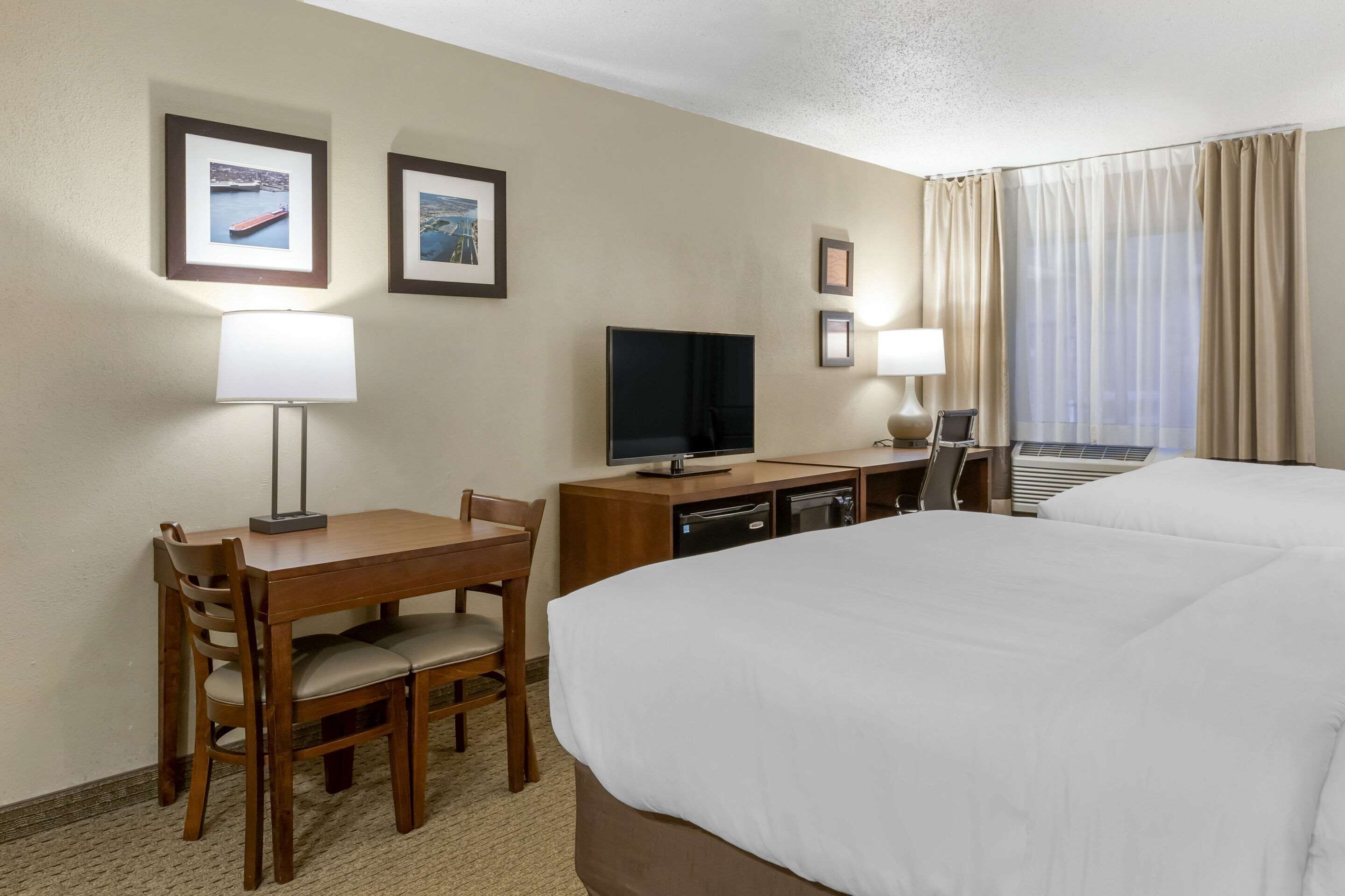 Une chambre d’hôtel avec un grand lit, un bureau avec un téléviseur à écran plat, deux chaises, une petite table et une fenêtre avec des rideaux.