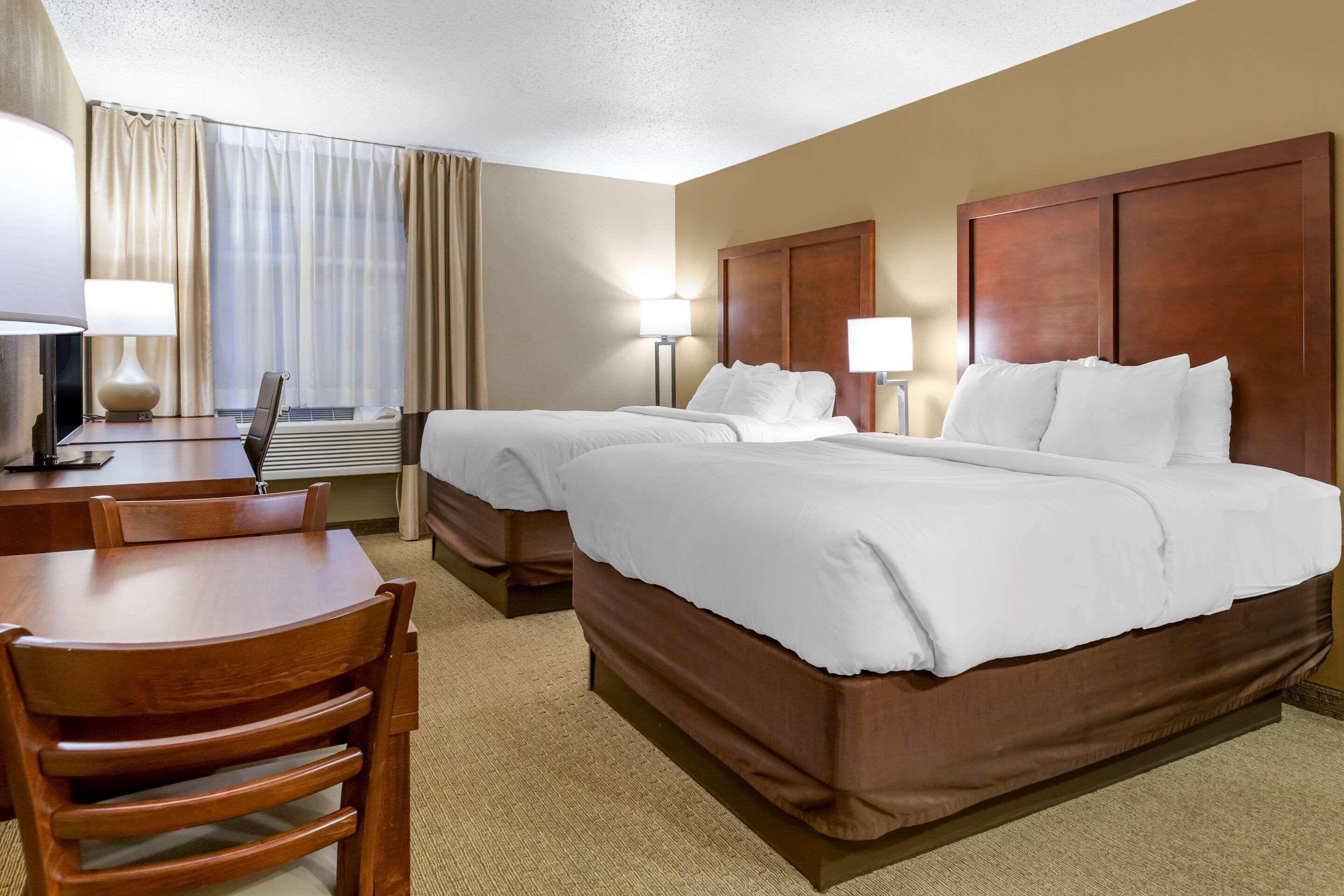 A hotel room with two beds, a desk, a chair, and a lamp.