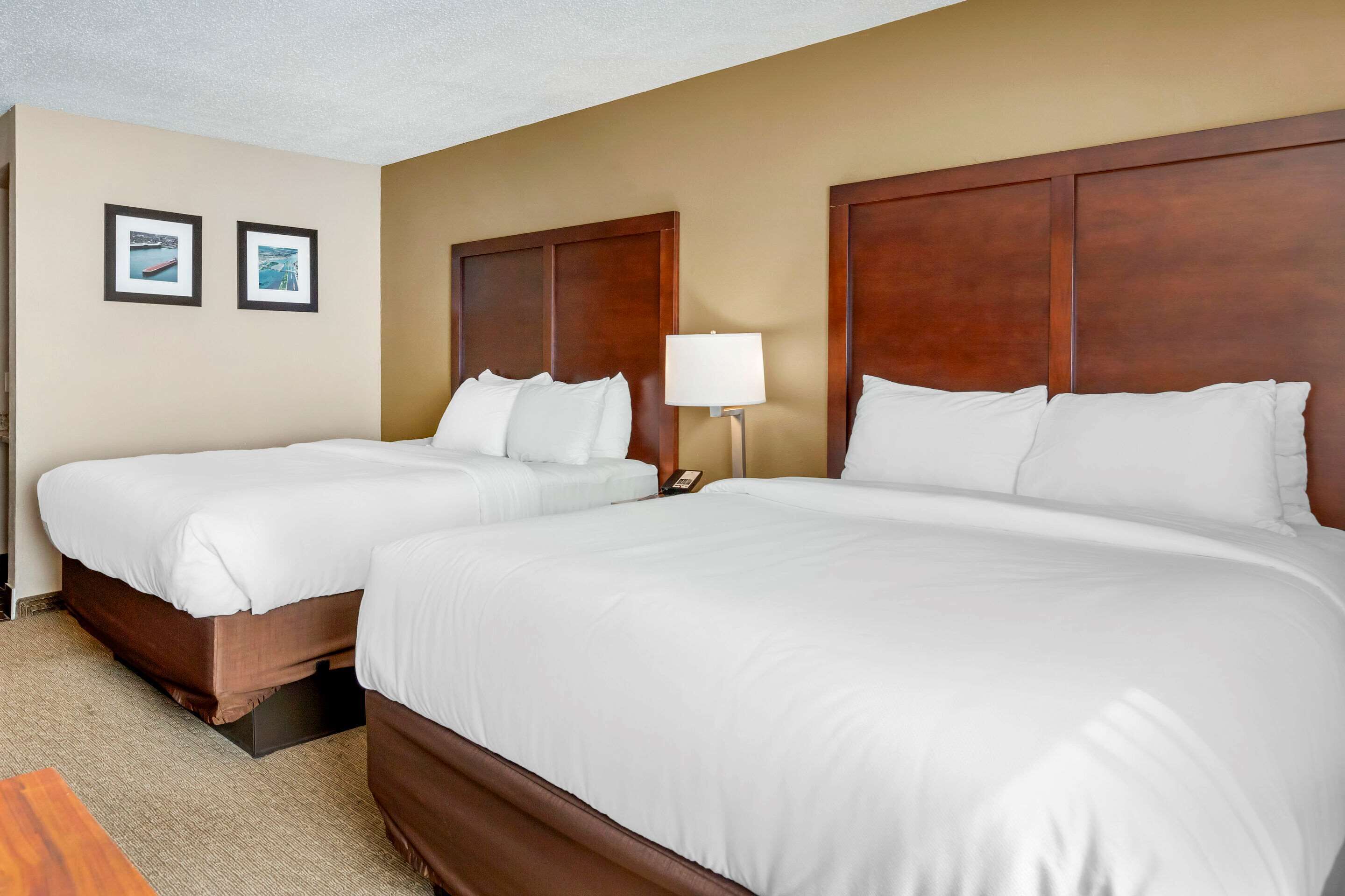 A hotel room with two single beds, a wooden headboard, a nightstand with a lamp, and two framed pictures on the wall.