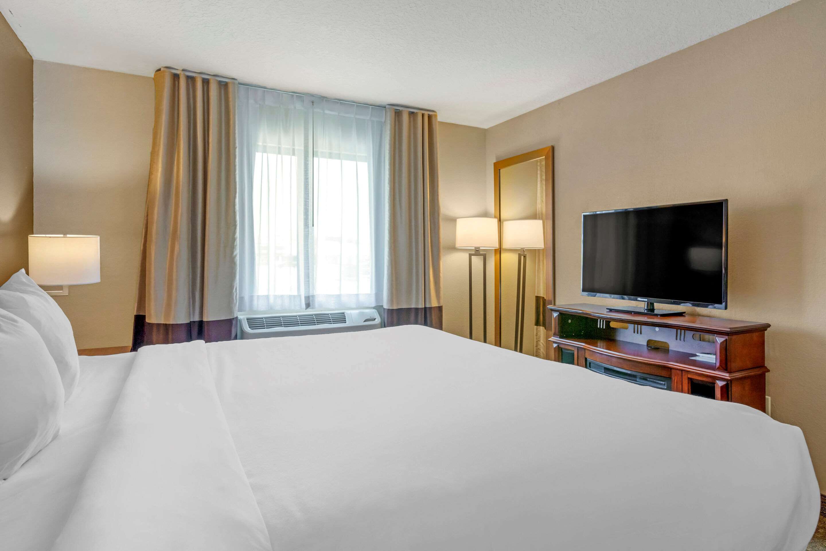A hotel room with a large bed, a flat-screen TV on a wooden stand, a mirror, and a window with curtains.