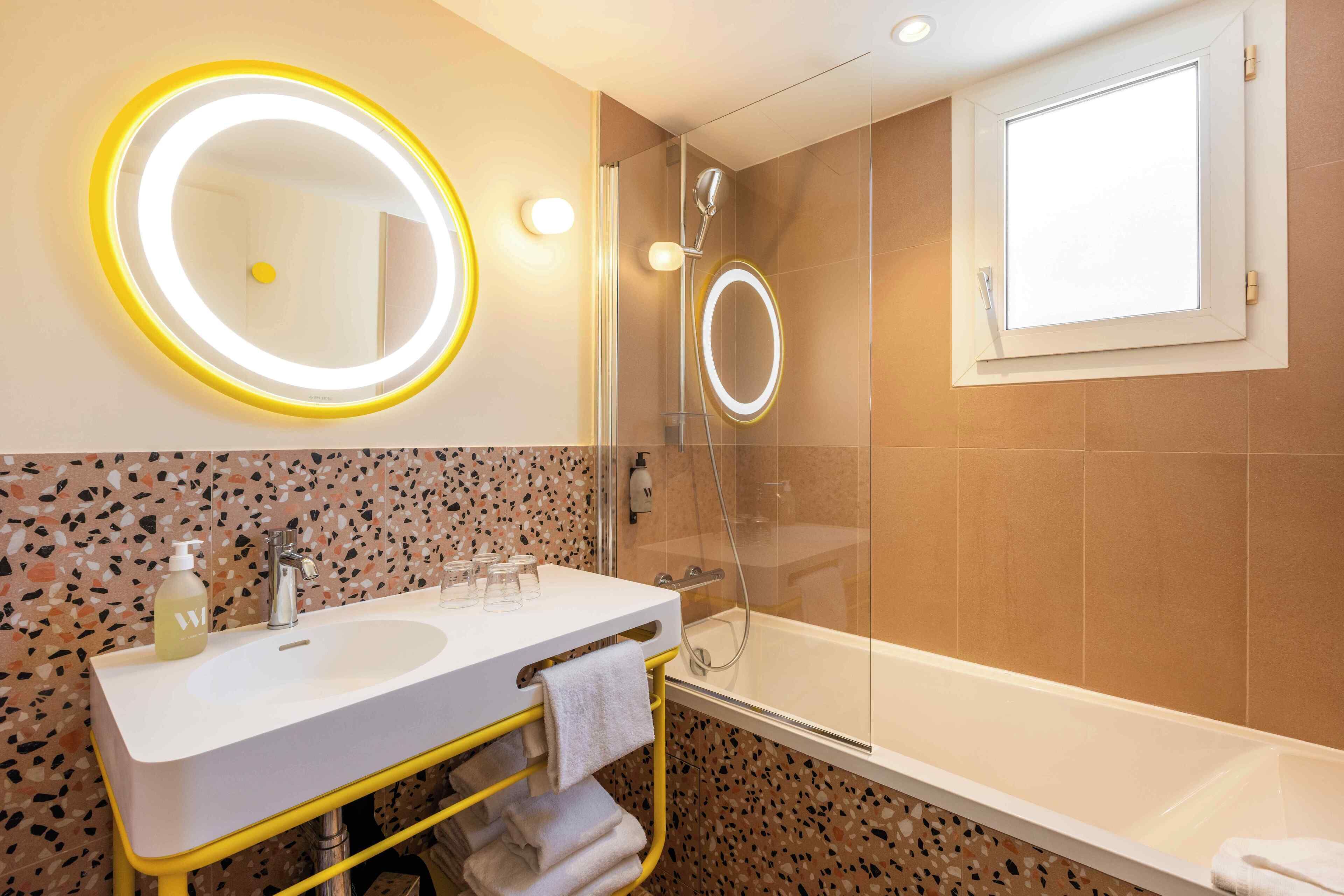 Une salle de bain avec un lavabo blanc, un miroir éclairé par une lumière jaune, une douche et une fenêtre.