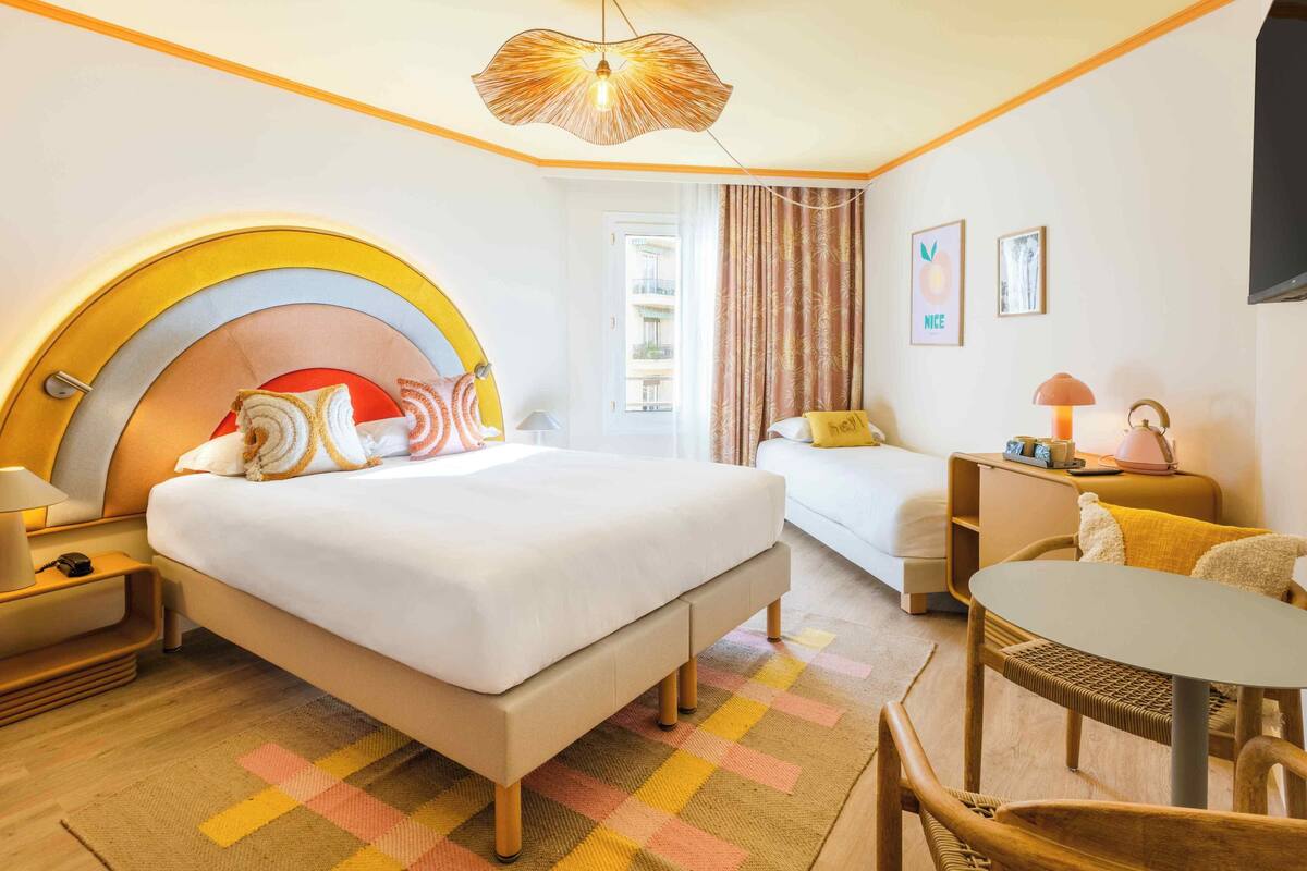 A hotel room with a bed, a desk, a chair, a lamp, and a view of the outside.