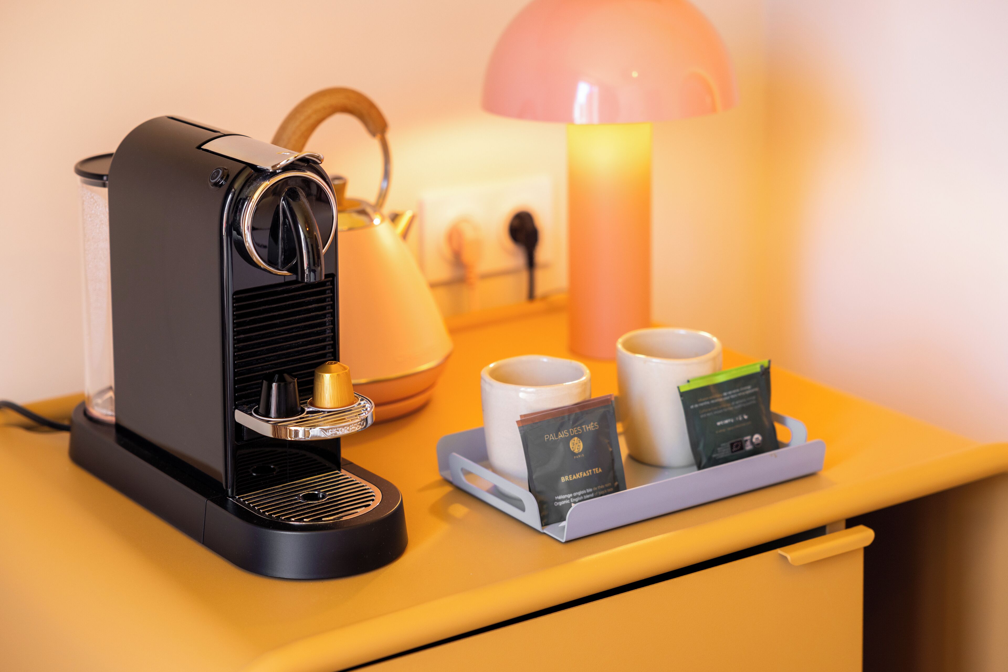 A coffee machine with a tray of cups and a tea packet on a yellow table.