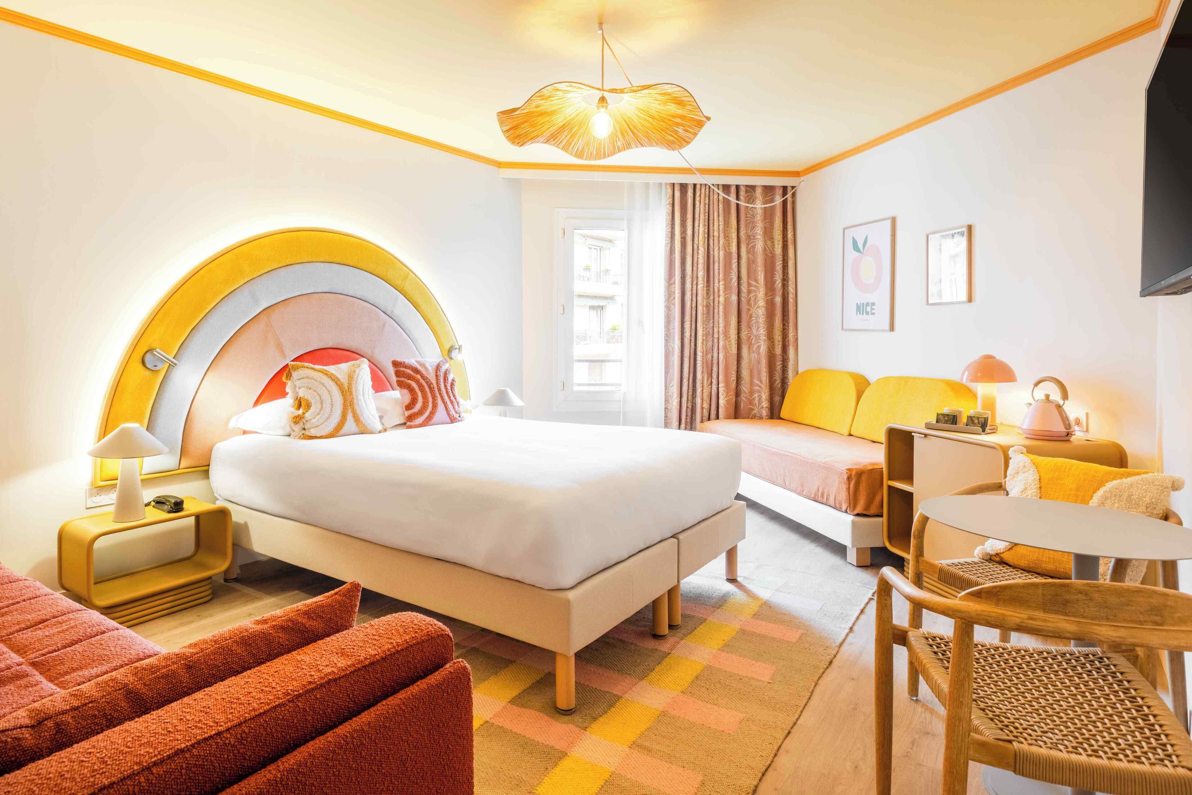 Habitación de hotel con cama, sofá, una mesita, y una silla. Se ve una ventana con cortinas, una lámpara en la mesita de noche, y un televisor instalado en la pared.