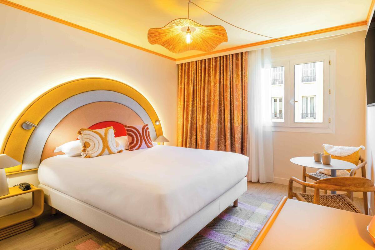 A hotel room with a large bed, a round headboard, a small table with chairs, a window with curtains, and a wall-mounted TV.