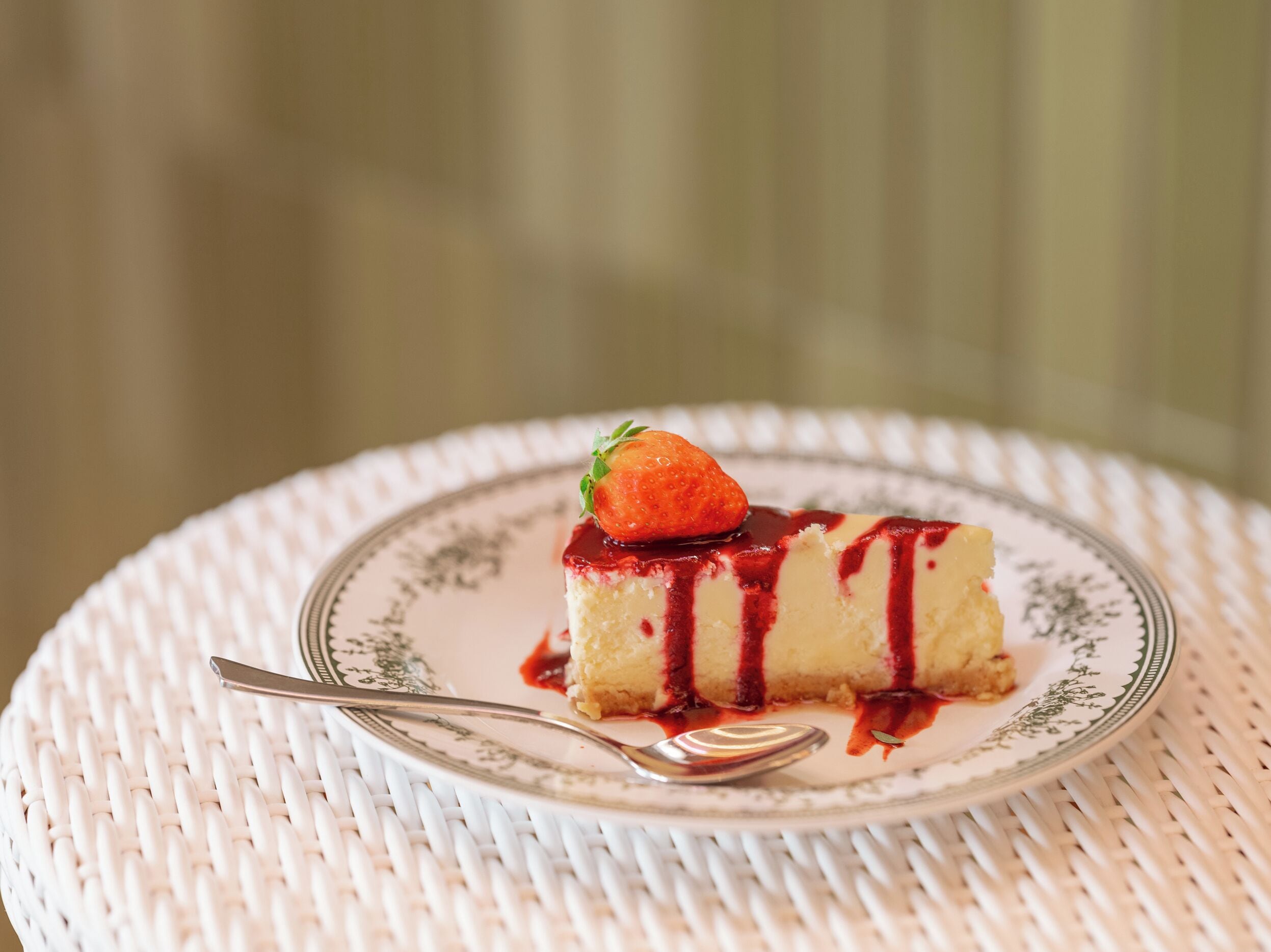 Una fetta di cheesecake su un piatto, con una fragola sopra e irrorata da salsa rossa, su un tavolo bianco intrecciato.