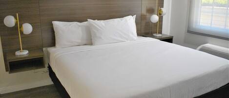 A modern hotel room with a large bed, white linens, a wooden headboard, bedside tables with lamps, and a window with blinds.