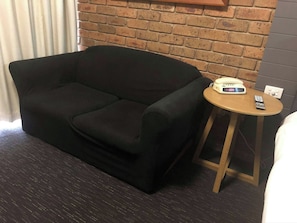 A dark-colored sofa in a room with a brick wall and a small wooden table with a clock and remote control. - Best Western Travellers Rest Motor Inn (Swan Hill)