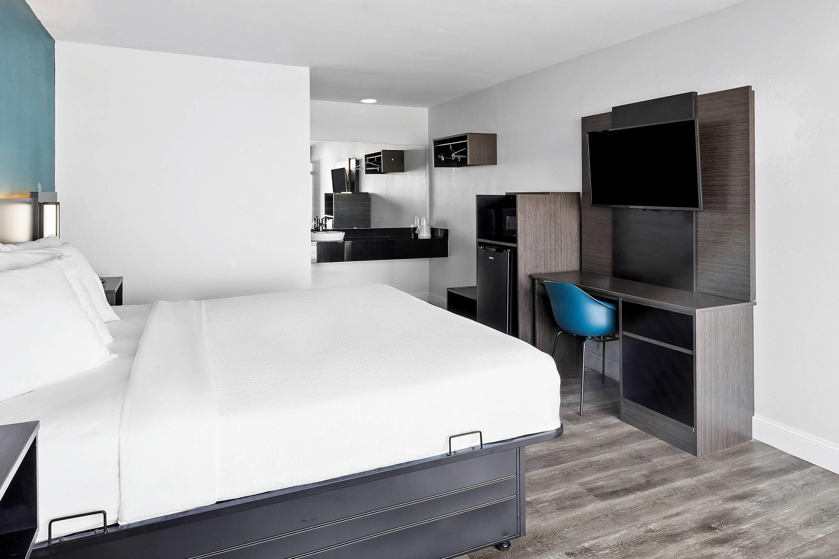 A modern hotel room with a large bed, a desk with a flat-screen TV, a built-in wardrobe, and a seating area with a blue chair.