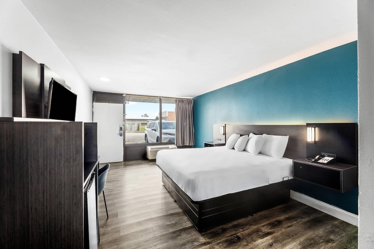 A modern hotel room with a large bed, a desk, a chair, a TV, and a window with a view of the outside.