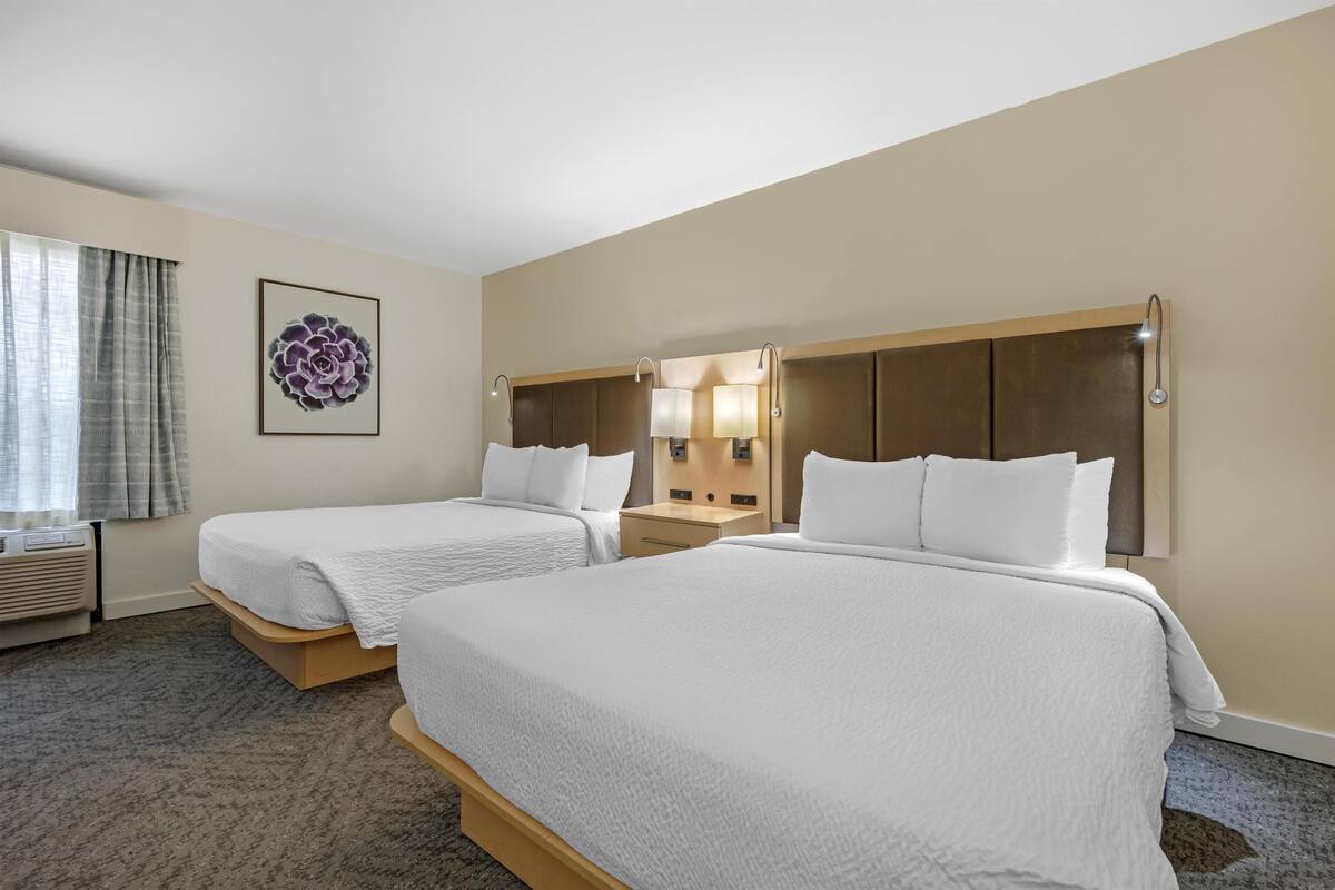 A hotel room with two beds, a nightstand, a lamp, and a wall-mounted artwork.