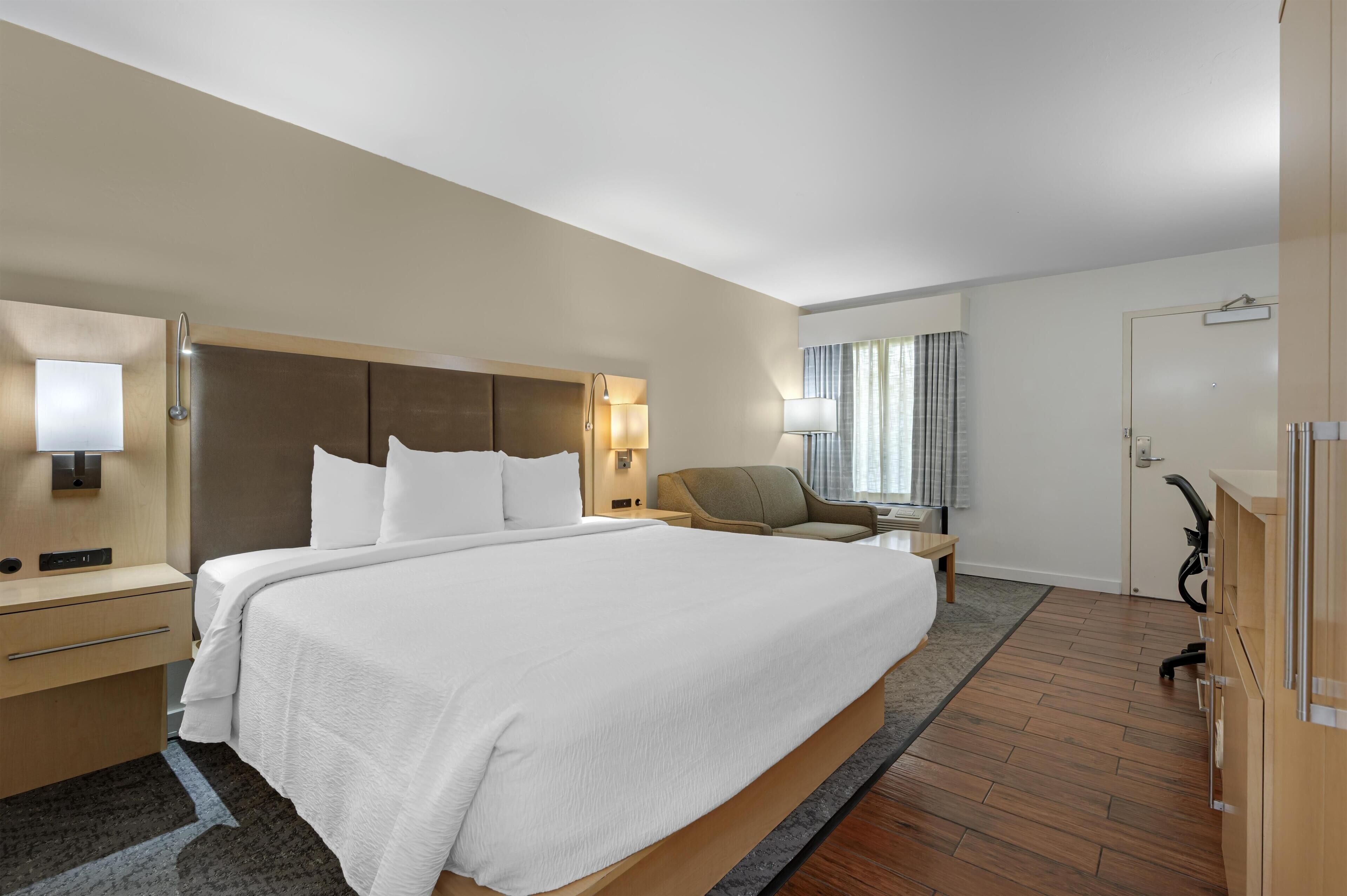 A modern hotel room with a large bed, bedside tables, a chair, and a desk.