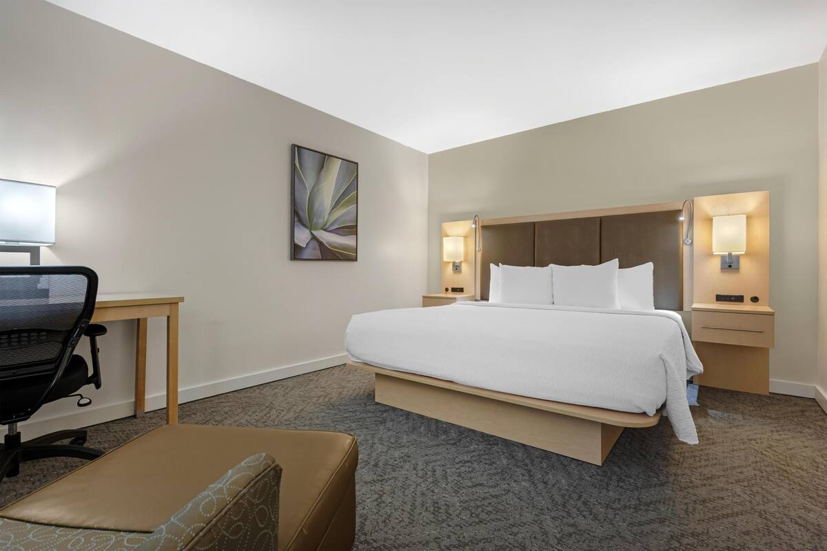 A hotel room with a large bed, a desk with a chair, a small table, and a wall-mounted lamp.