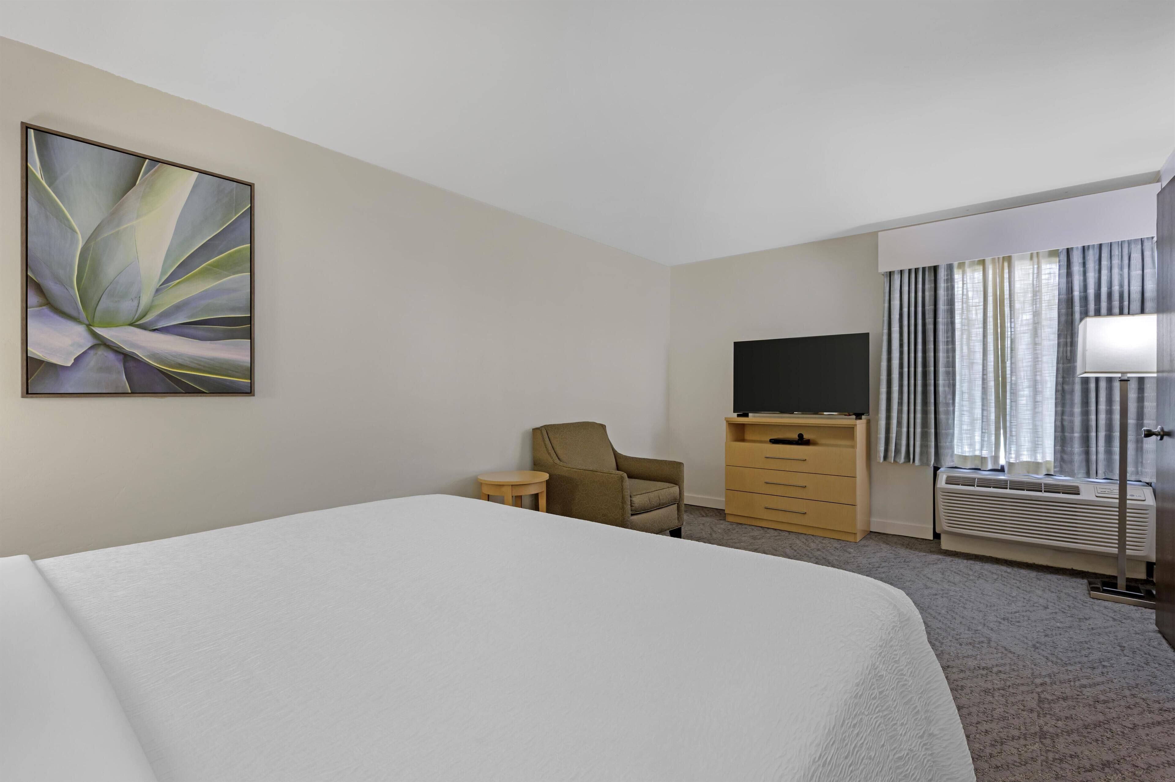 A hotel room with a large bed, a chair, a TV, a dresser, and a window with curtains.
